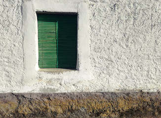 Rustic window of an agricultural village. Old construction facade. Green shutter of old wood. The rural Spain. White paint on stone receiving the sun's rays. Minimalist natural contrast of the window.