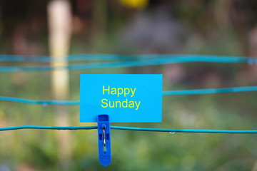 Conceptual photo: Blue clothes peg with blue note written "Happy Sunday". Selective focus