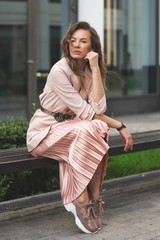 Young European in a jacket, pleated skirt, coral-colored sneakers resting on a bench in the street near the business center. Horizontal portrait of a beautiful girl.