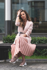 Fototapeta premium girl sitting on a bench on the street near the building with shop windows. Coral, pink jacket, pleated skirt, sneakers. Young beautiful European.