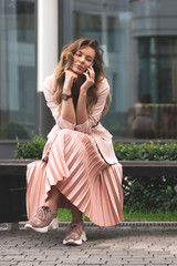 fashion girl sitting on a bench and talking on the phone. Lifestyle summer. Relaxed posing. Women's coral-colored clothing - a jacket, pleated skirt, sneakers, black blouse.