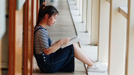 Young student girl with ponytail sitting on floor in library and leaning on shelf while reading a book on her legs.