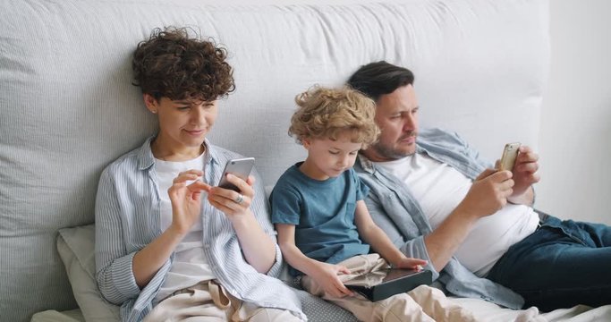 Little boy son is playing game on tablet touching screen while parents are using smartphones in bed at home. Modern lifestyle, family and device addiction concept.