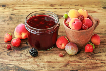 Jar of strawberry jam among summer and autumn fruits on a wooden table.