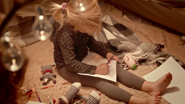 Sisters draw with pencils in a decorative tent in the room at night.