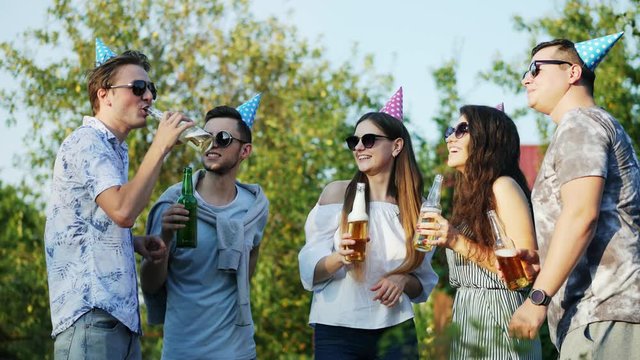 Group of friends drinking beer from bottle on nature, young people dancing on summer party, wearing birthday caps