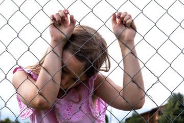 Sad girl behind metal wire mesh fence. Abandoned upset and lonely child outdoor. Depressed preteen...