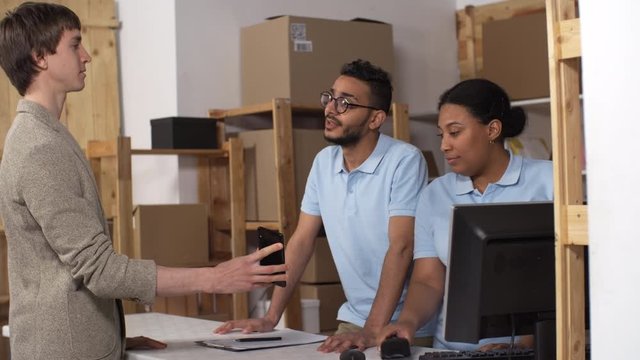 Medium shot of two friendly mixed race clerks working in post office branch, customer walking in, showing tracking code on smartphone, collecting parcel, and woman scanning and typing on computer