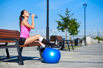 Fitness woman with bottle and rubber fit ball. Female drinking water after training on bench in summer park