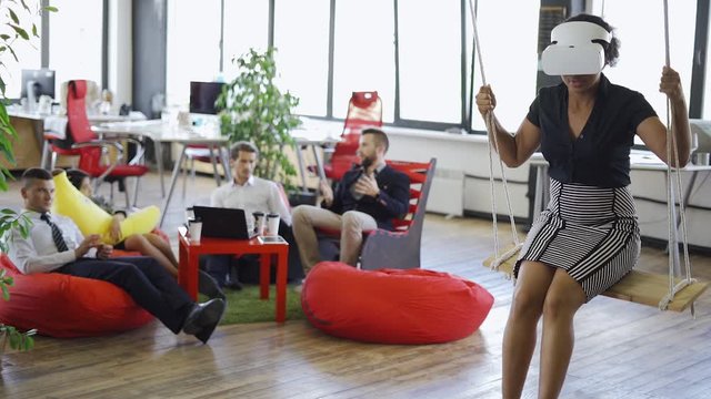 Young african woman in futuristic virtual reality glasses riding on a swing, modern office and workers on a blurry backround.
