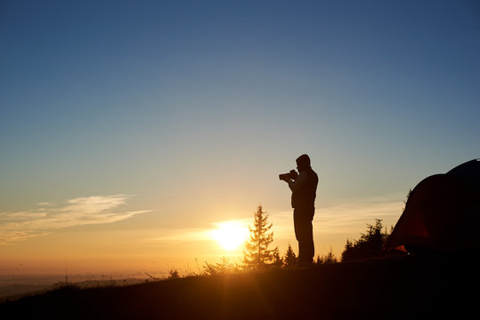 Silhouette of young male photographer with photo camera standing beside tourist tent on the top of mountain in the morning at sunrise. On background beautiful view of rising sun and blue sky.