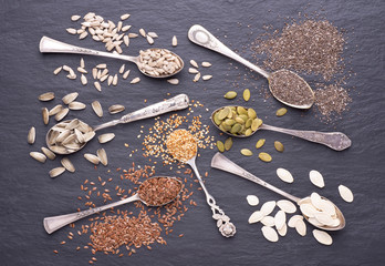 Various seeds - sesame, flax seed, sunflower seeds, pumpkin seed, chia in spoons on a black stone background. Top view