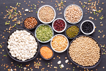 Legumes, lentils, chickpeas and beans assortment in various bowls on black stone background, top view