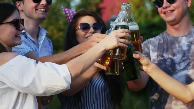 Close-up of group of friends clink with beer bottles on summer party while celebrate birthday, in holiday caps