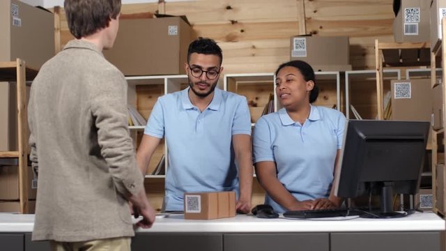 Thigh-up shot of Afro-American female and Arab male postal employees in uniform blue polos working behind counter and greeting entering customer, who is signing on tablet, taking parcel and leaving