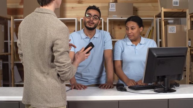 Medium shot of customer walking into post office branch and showing tracking number on smartphone to two polite multiethnic clerks, who are scanning it, giving him package, smiling and saying goodbye