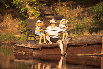 Cute little girls and their granddad are on fishing at the lake or river. Resting on pier near by water and forest in sunset time of summer day. Concept of family, recreation, childhood, nature.