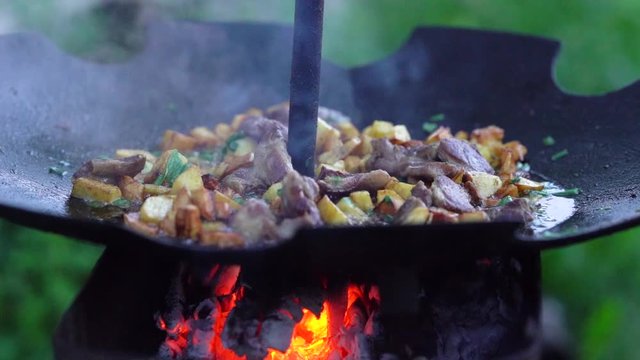 Close-up scene of a potatoes with meat which are roasting on a barbecue pan on a fire. There is a picnic in a forest.