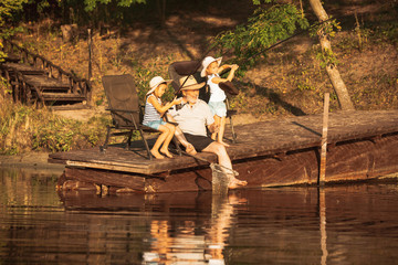Cute little girls and their granddad are on fishing at the lake or river. Resting on pier near by water and forest in sunset time of summer day. Concept of family, recreation, childhood, nature.