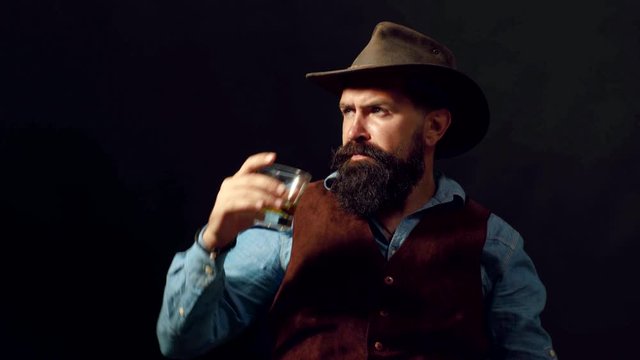 Cowboy with cowboy hat holding whisky cocktail in glass. Hipster with beard and mustache in suit drinks alcohol after working day.