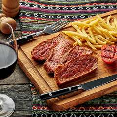 Steak with French fries and grilled cherry tomatoes on a cutting board. Classic meat meal with red wine on an ethnic tablecloth.