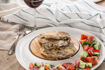 Homemade lamb schnitzel with fresh vegetable salad on the side and glass of red wine on tablecloth