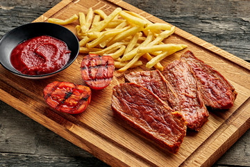 Steak with French fries, grilled cherry tomatoes, sauce and red wine on a rustic wooden table. Wholesome American lunch served on a cutting board. Close-up shot.