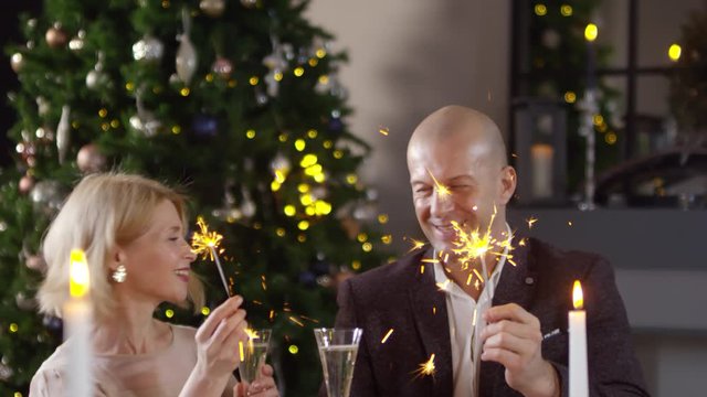 Tilting medium shot of 40-something Caucasian man and woman celebrating New Year together at home with romantic dinner and champagne, lighting up sparklers, looking at each other and touching glasses