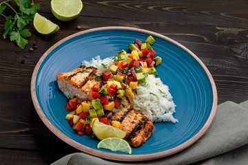 Salmon fillet cooked on grill with exotic sauce made of mango, avocado and pepper on blue plate. Delicious fish meal with jasmine rice on a table.