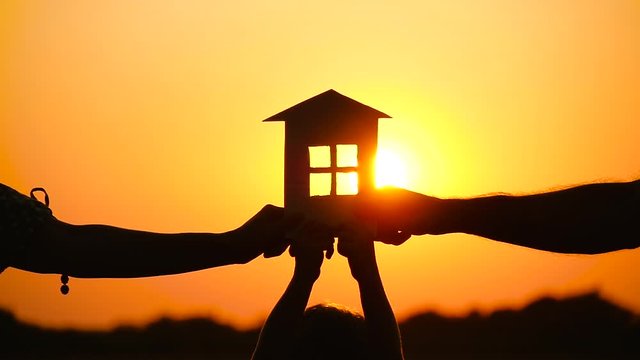 A happy family keeps a paper house on the background of a beautiful sunset. The concept of family relationships, love and real estate.