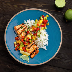 Grilled salmon fillet with fruit sauce made of mango, bell pepper, onion and avocado with basmati rice garnish on a blue plate. Delicious seafood meal. Top view shot above.