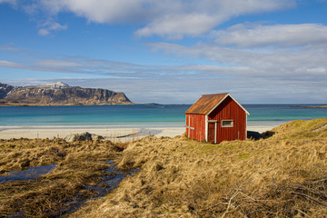 Fototapeta na wymiar A typical red nordic hut at the seaside. Amazing and beautiful sunlit beach with mountain range in the background.