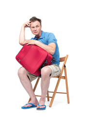 Man in a blue T-shirt and white shorts sits on a chair near a red suitcase, waiting. Isolated on white background
