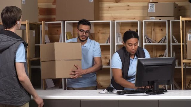 Thigh-up Shot Of Two Multiethnic Post Office Employees Standing Behind Desk, Caucasian Courier With Earpiece Walking In, Arab Worker Taking Out From Rack And Black Clerk Scanning Two Large Packages