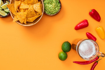 top view of nachos, guacamole, beer, limes and peppers on orange background with copy space