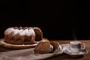 Delicious chocolate sponge cake on a wooden plate and two slices of the cake on a napkin next to a cup of hot coffee. Rich snack for a cold fall or winter day. Black background. Space for text