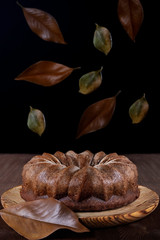 Delicious chocolate sponge cake on a wooden plate and table on which dry leaves are falling as in autumn. Vegetarian and healthy food for the autumn season. Black background