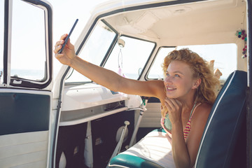 Woman taking selfie with mobile phone while lying on seat in camper van at beach