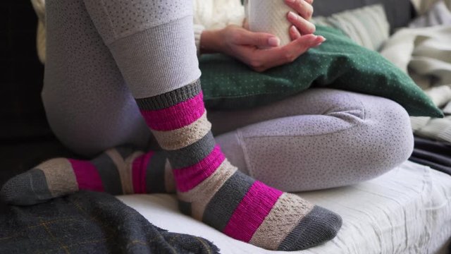Pink and brown warm woolen socks close-up - a woman is sitting with a white cup of hot drink on the sofa with a plaid and pillows. Lagom or Hygge concept - comfort and simplicity