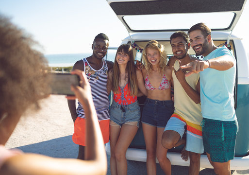 Woman taking pictures of her friends with mobile phone near camper van at beach 