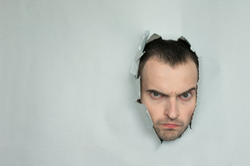 Angry guy making hole in paper, looking at the camera, portrait, blue background, copy of the space
