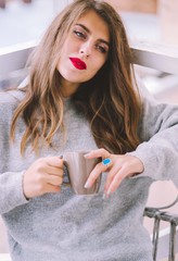 Relaxed woman with cup of coffee or tea. Stylish girl dressed in a gray sweater and silk skirt resting on the balcony.