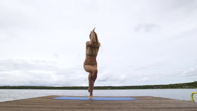 Fit slim woman practicing yoga exercises wearing mini swimsuit bikini at lake with clouds - Yoga meditation and wellness lifestyle concept - 16mm wide full low shot
