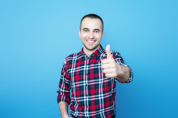 Attractive guy in plaid shirt smiling looking at camera, standing and showing thumb up, front view, blue background