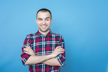 Attractive guy in plaid shirt smiling looking at camera, standing and having his arms crossed, front view, blue background