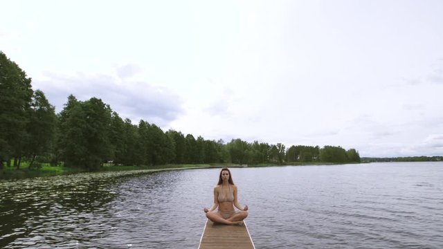 Slow gimbal meditating pan: Fit slim woman practicing yoga exercises wearing mini swimsuit bikini at lake with clouds - Yoga meditation and wellness lifestyle concept - Full sitting shot