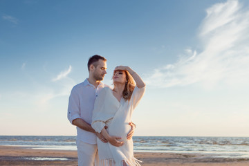 Romantic walk on the beach, waiting for a baby. Happy young couple hugging tummy, spending time by the sea.