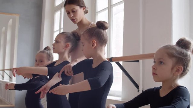 Medium shot of young Caucasian female teacher going along studio and showing little ballerinas how to hold hands in right position