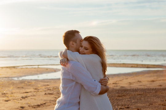 Love, sunset, romance. A young couple in long white soft clothes hugging against the backdrop of a calm sea, sunset.