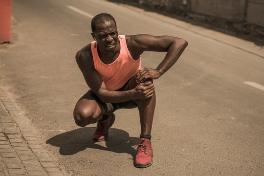 young athletic and fit black African American runner in pain holding his knee after suffering medical problem with injuried tendon or ligament during urban running workout